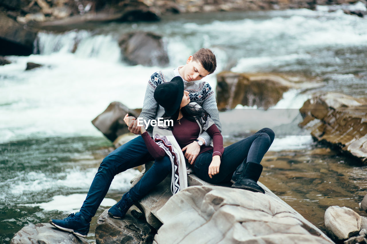 PEOPLE SITTING ON ROCK BY WATER