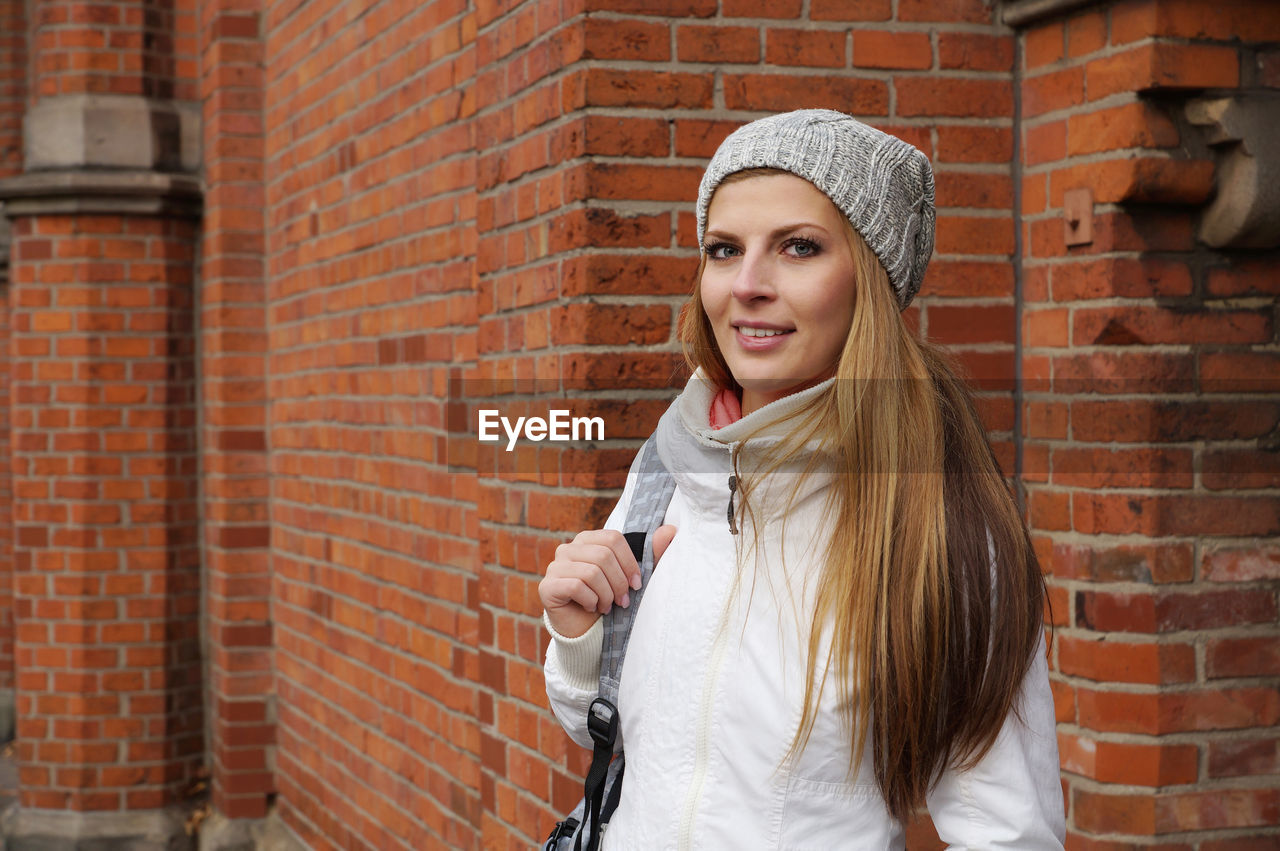 Portrait of smiling beautiful woman standing against brick wall during winter