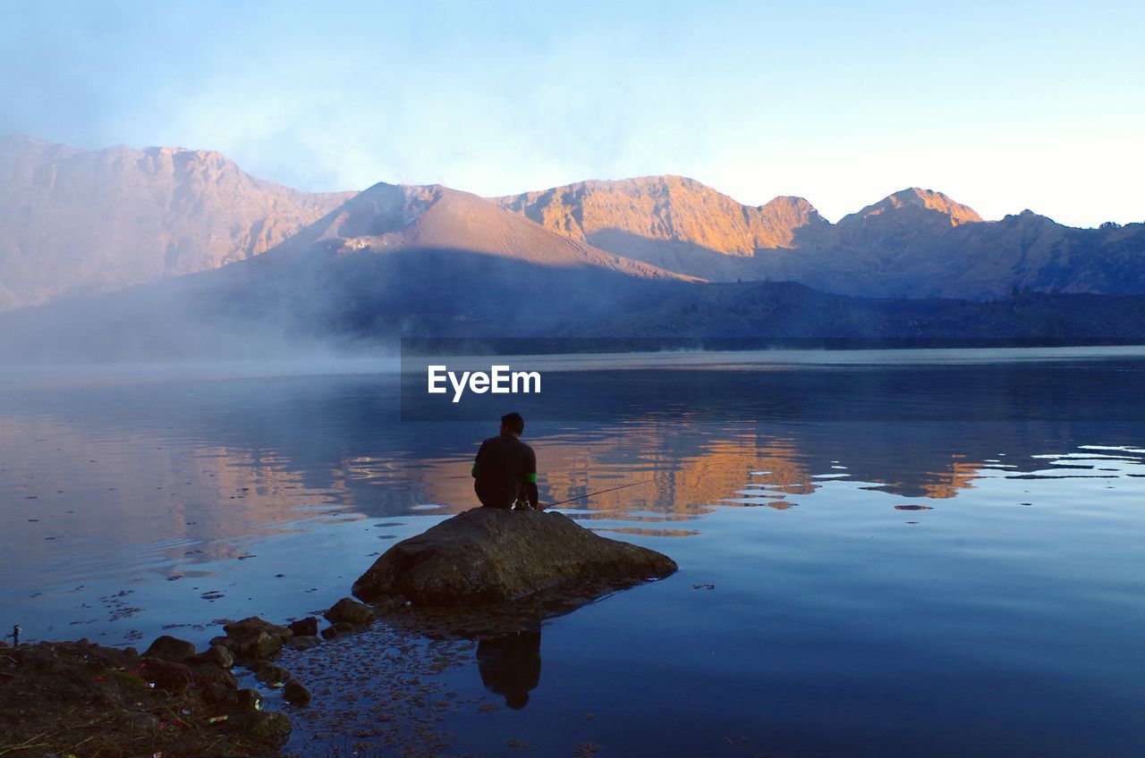REAR VIEW OF MAN LOOKING AT LAKE AGAINST MOUNTAIN RANGE