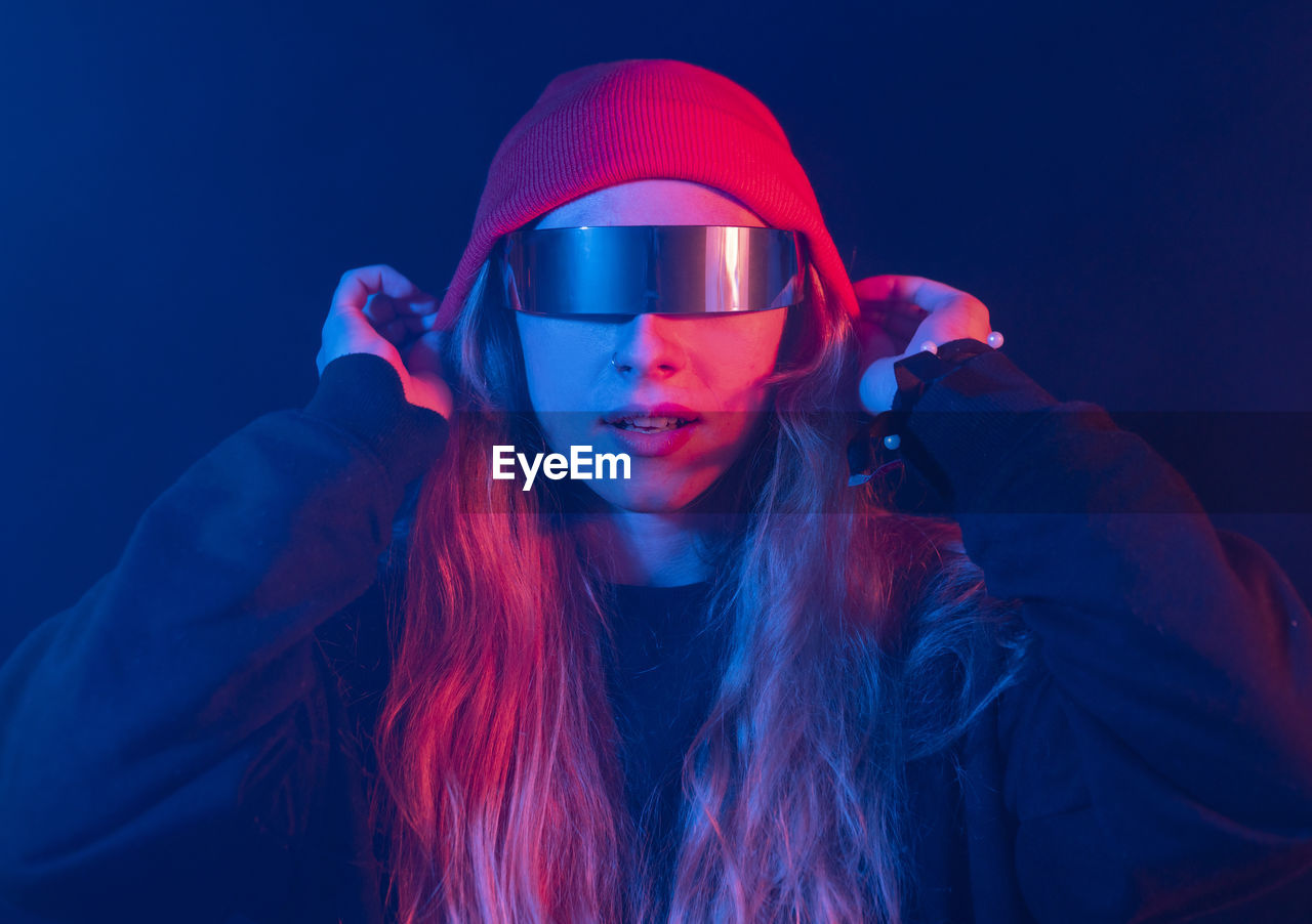 Trendy young female millennial with long blond hair in futuristic sunglasses adjusting hat while standing in dark room with neon illumination
