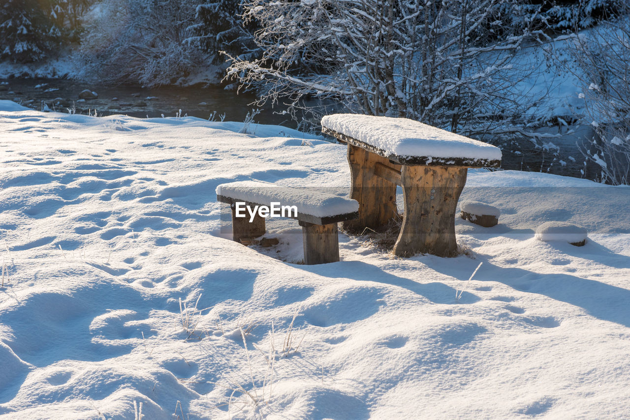 Seats on snow covered landscape by trees and water during winter