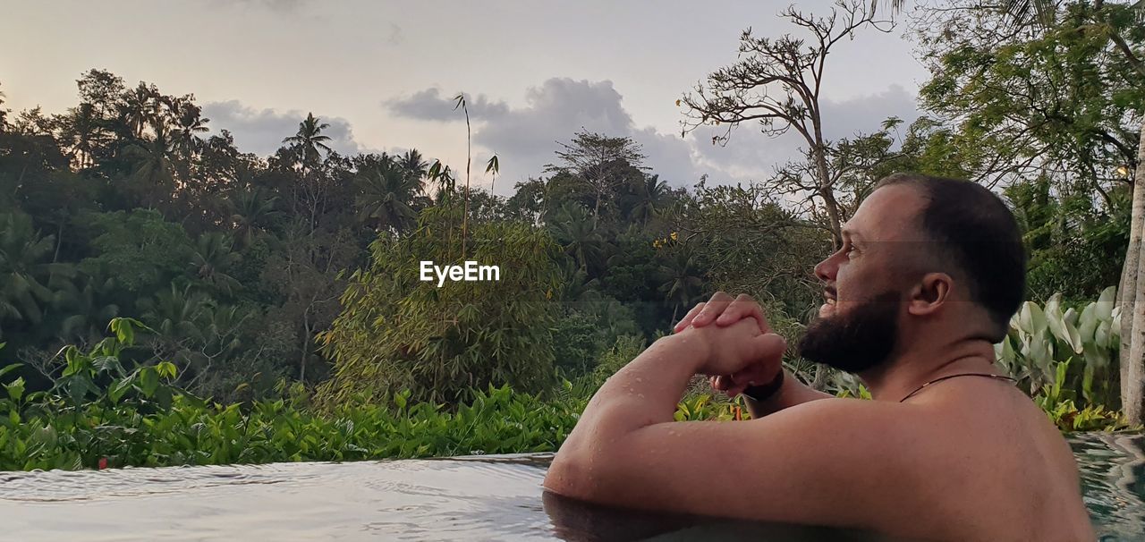 SIDE VIEW OF SHIRTLESS YOUNG MAN AGAINST PLANTS