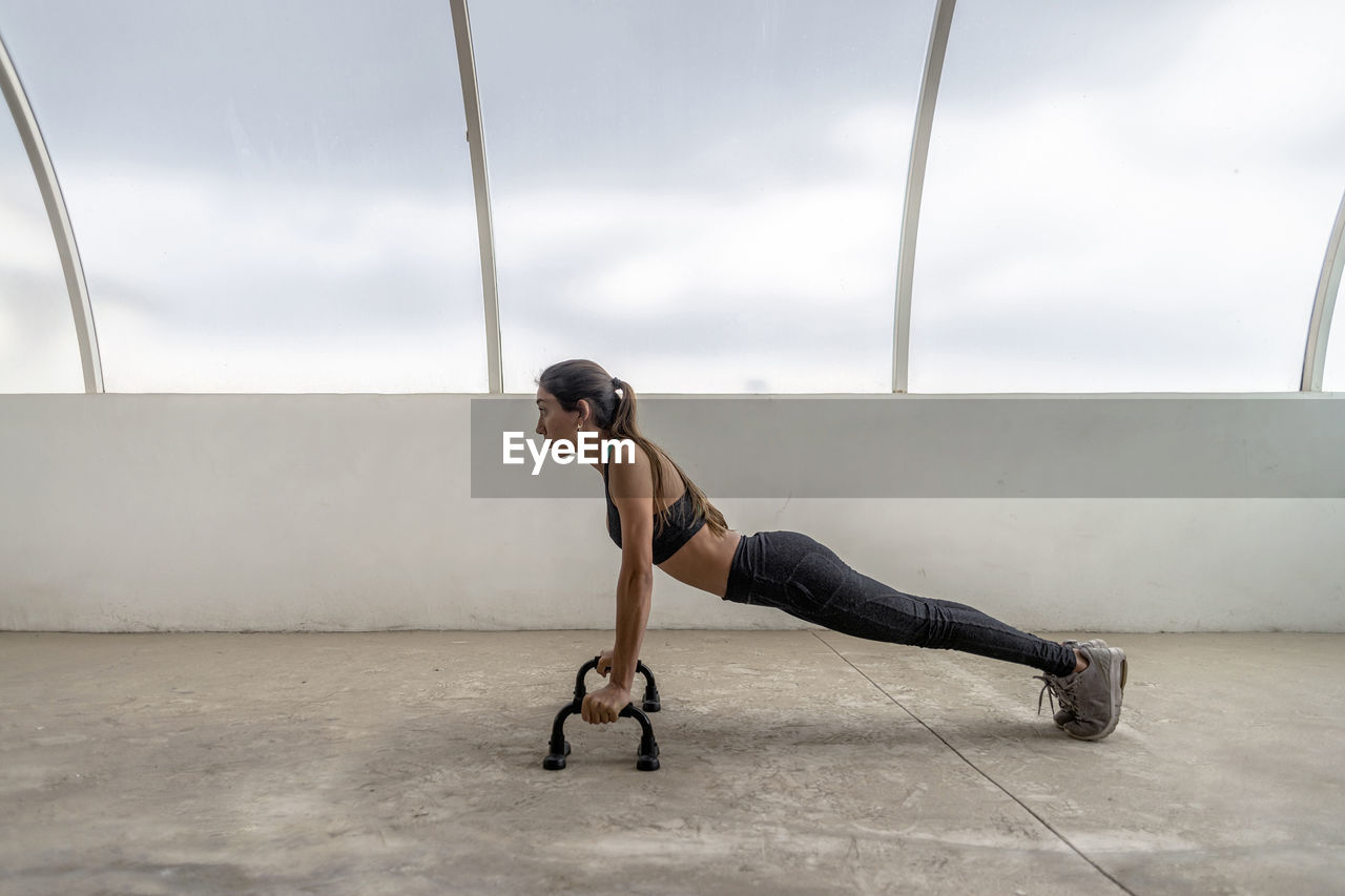 Side view of fit ethnic sportswoman in active wear exercising on push up bars while looking forward