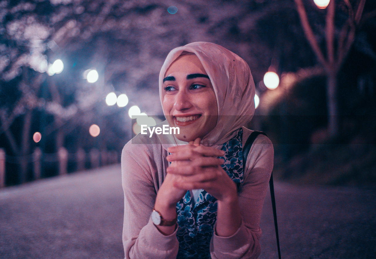 PORTRAIT OF SMILING YOUNG WOMAN AGAINST ILLUMINATED LIGHT