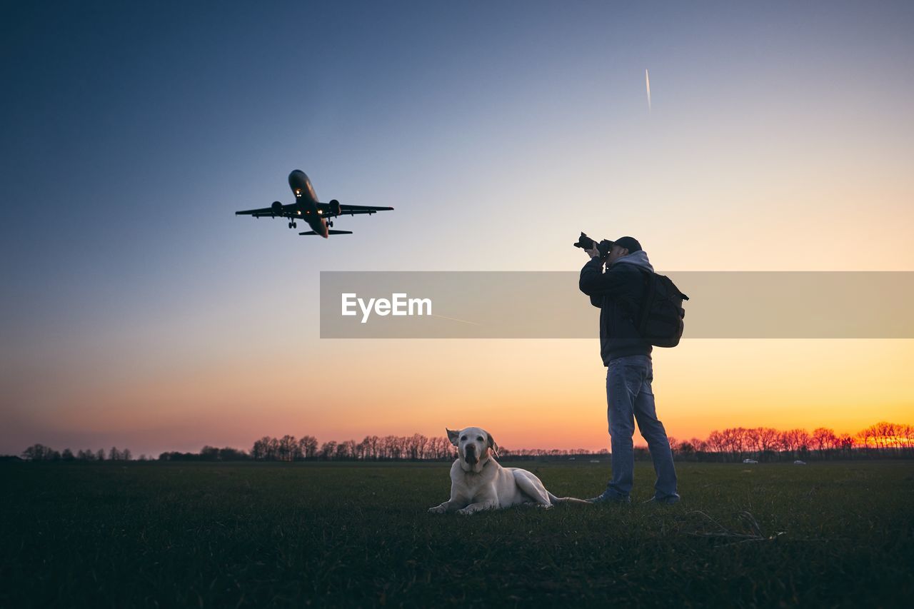 Mid adult man photographing airplane while standing by dog on field against sky during sunset