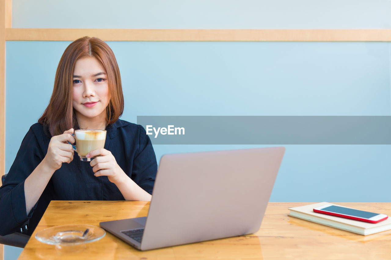 Portrait of businesswoman having coffee while using laptop at desk in office