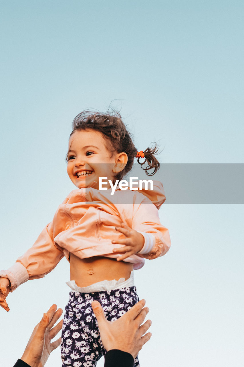 Cropped hands of father throwing cheerful daughter in air against clear sky