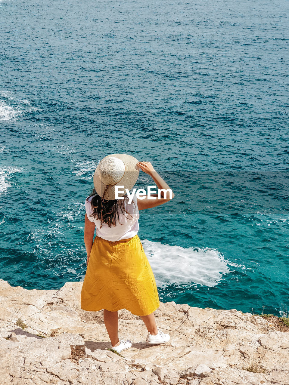 Young woman in yellow skirt standing on edge of cliff above sea.