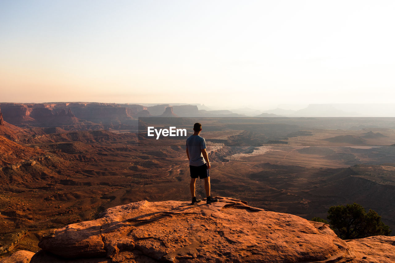 REAR VIEW OF MAN STANDING ON ROCK AGAINST MOUNTAIN