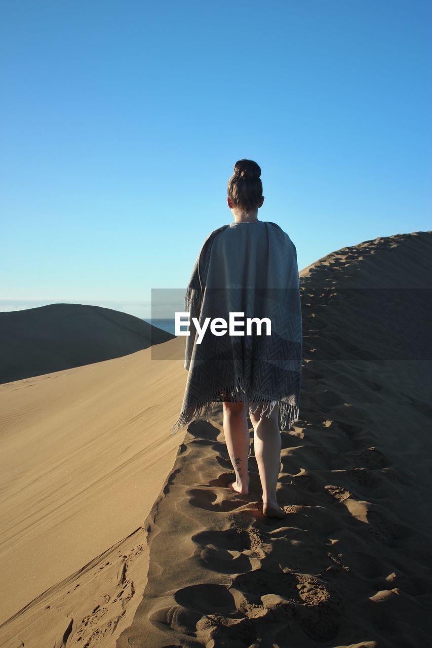 Rear view of woman with blanket walking on sand dune in desert against clear sky