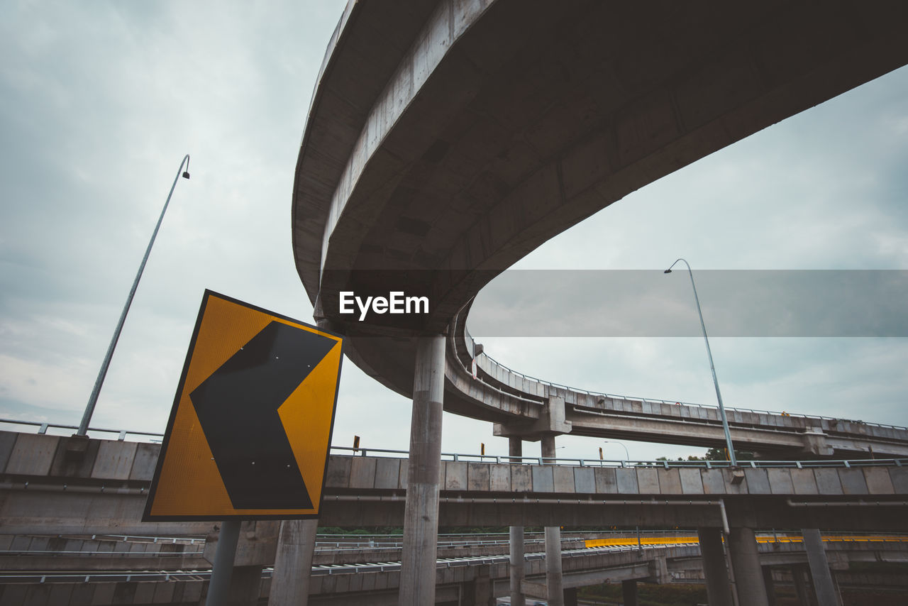 Low angle view of road sign below bridges against cloudy sky