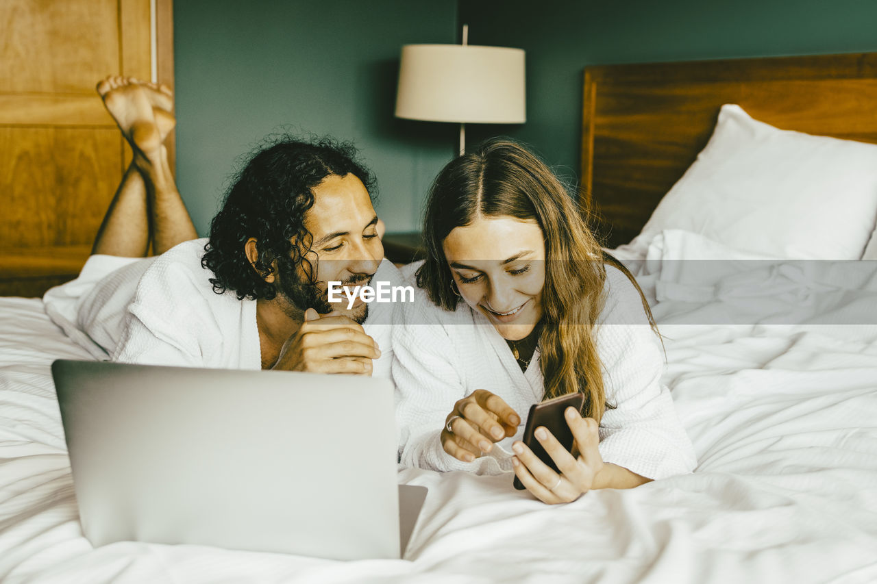 Couple in bathrobe with laptop using phone while lying on bed at hotel room