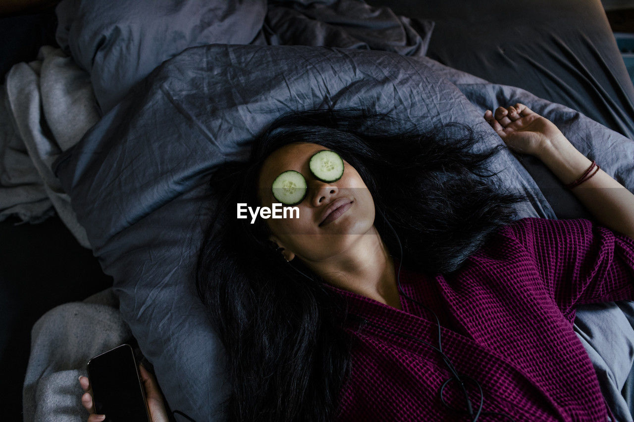 High angle view of woman lying on bed with cucumber slices on eyes