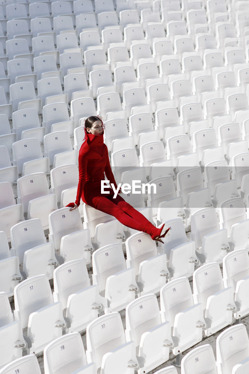 Full length of fashion model in red sitting on white chairs at stadium