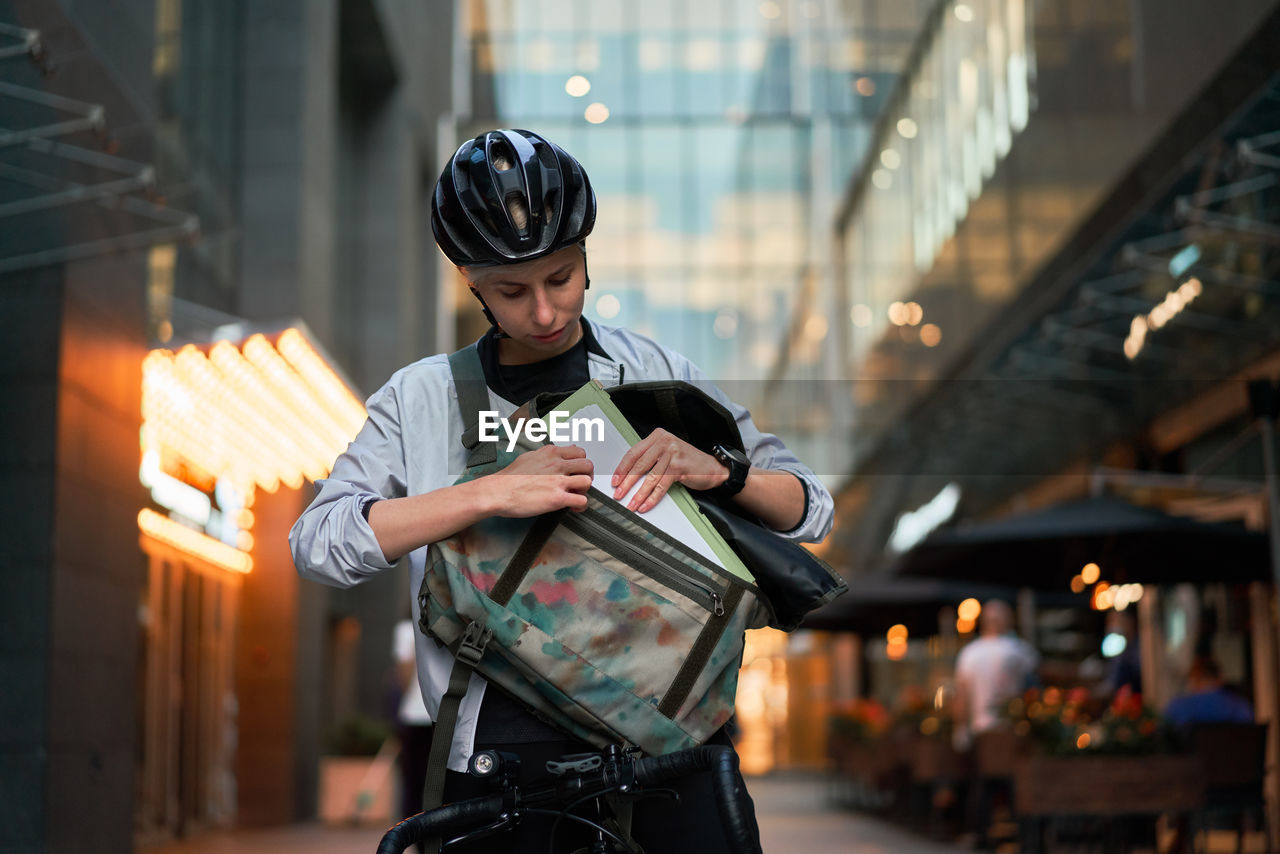 Close-up of woman in helmet on bicycle looking into bag near tall building with glass windows