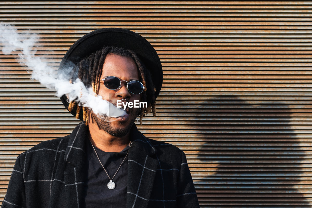 Modern african american man with dreadlocks wearing hat with sunglasses and exhaling smoke on street
