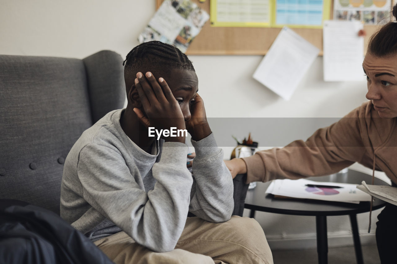 Non-binary mental health professional consoling sad male student sitting on chair in office