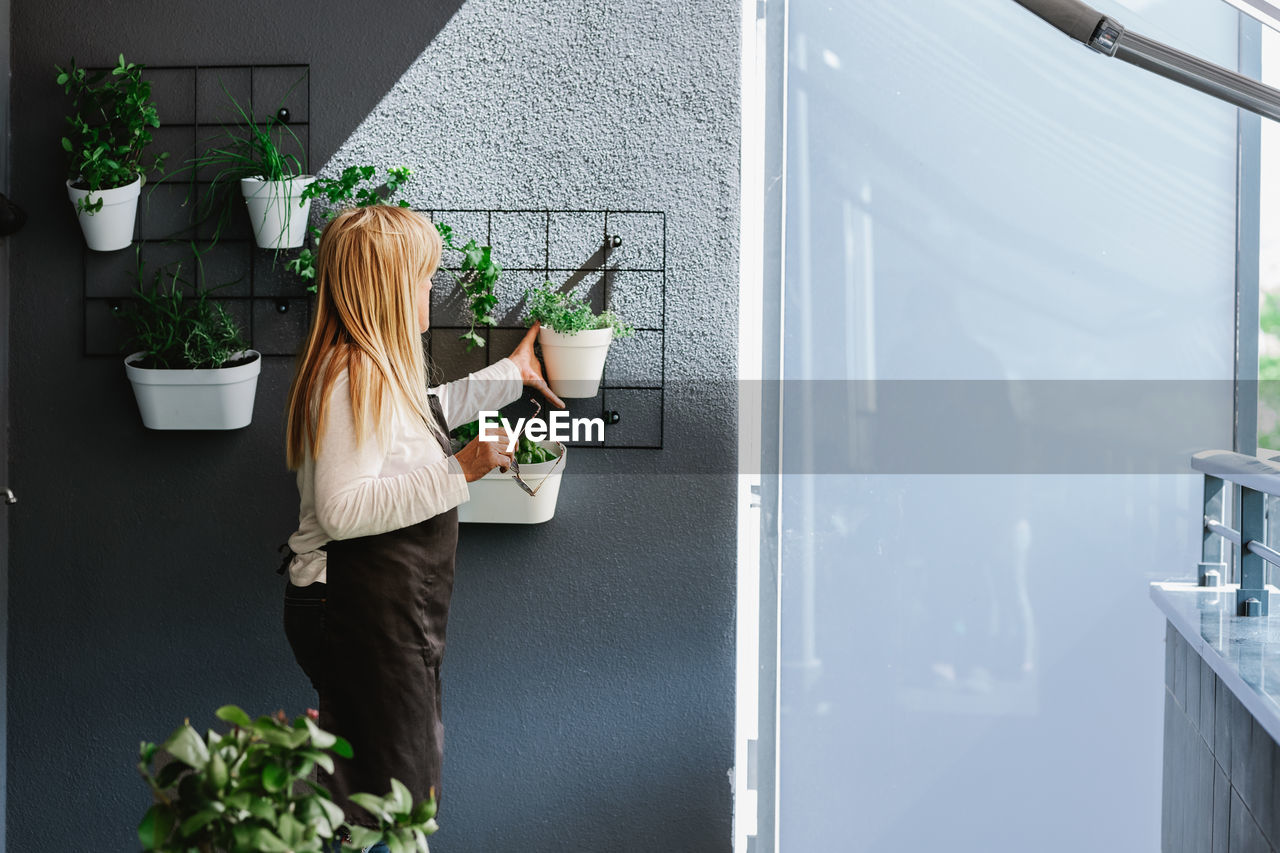 Side view of female hanging white flowerpot with green plant on gray wall with various flowers in light room