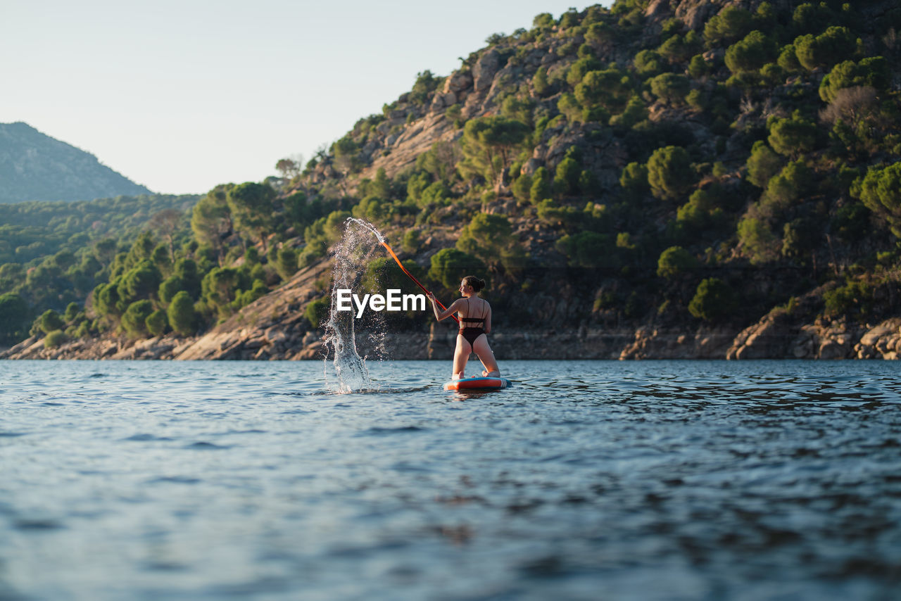 Full body of active female in swimwear floating on paddleboard in rippling lake against rock and shore with forest on summer day