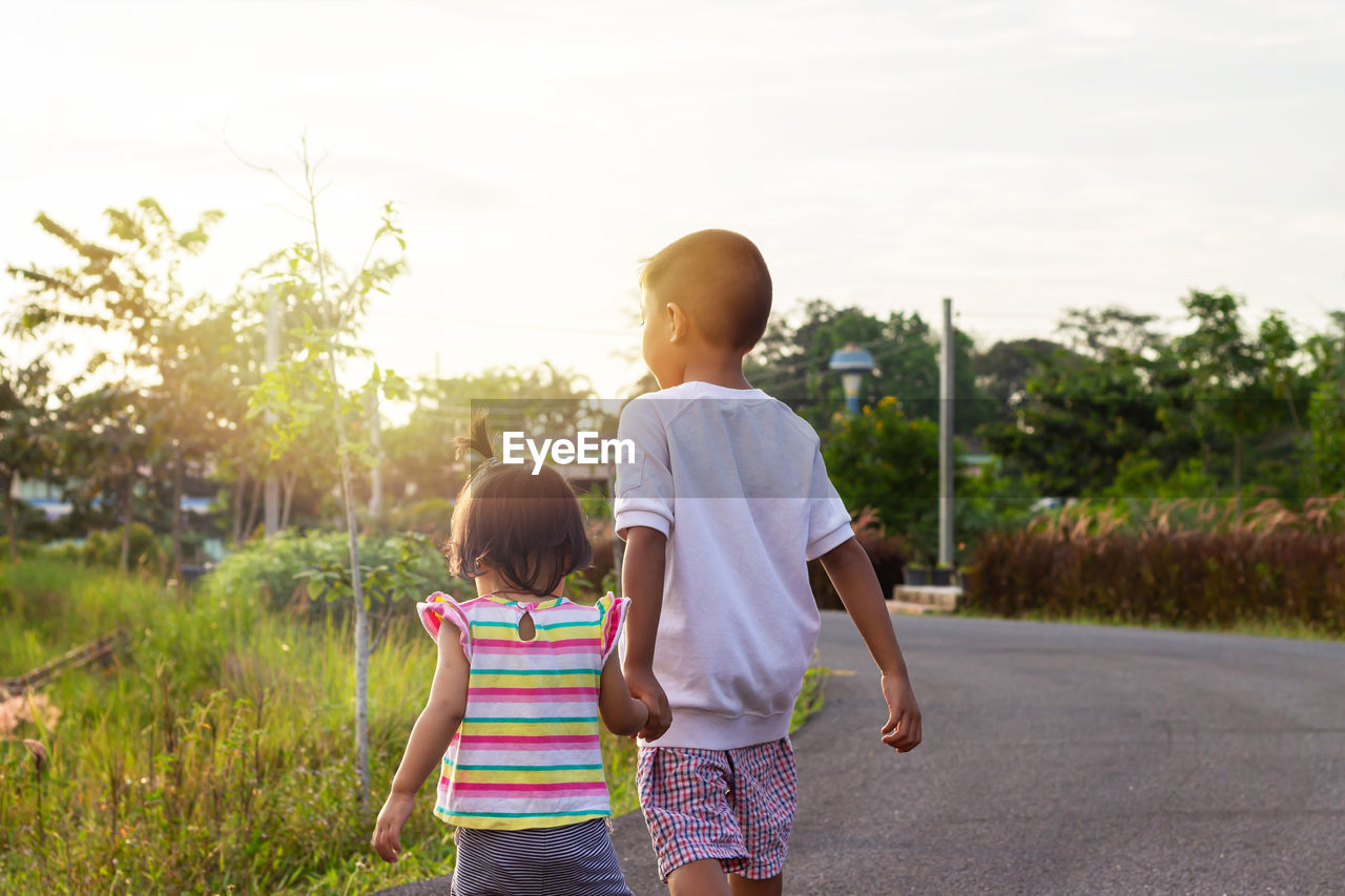 Rear view of siblings holding hands while walking on road against sky