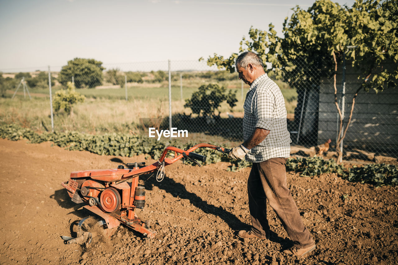 Senior male farmer plowing field with electric harrow on sunny day