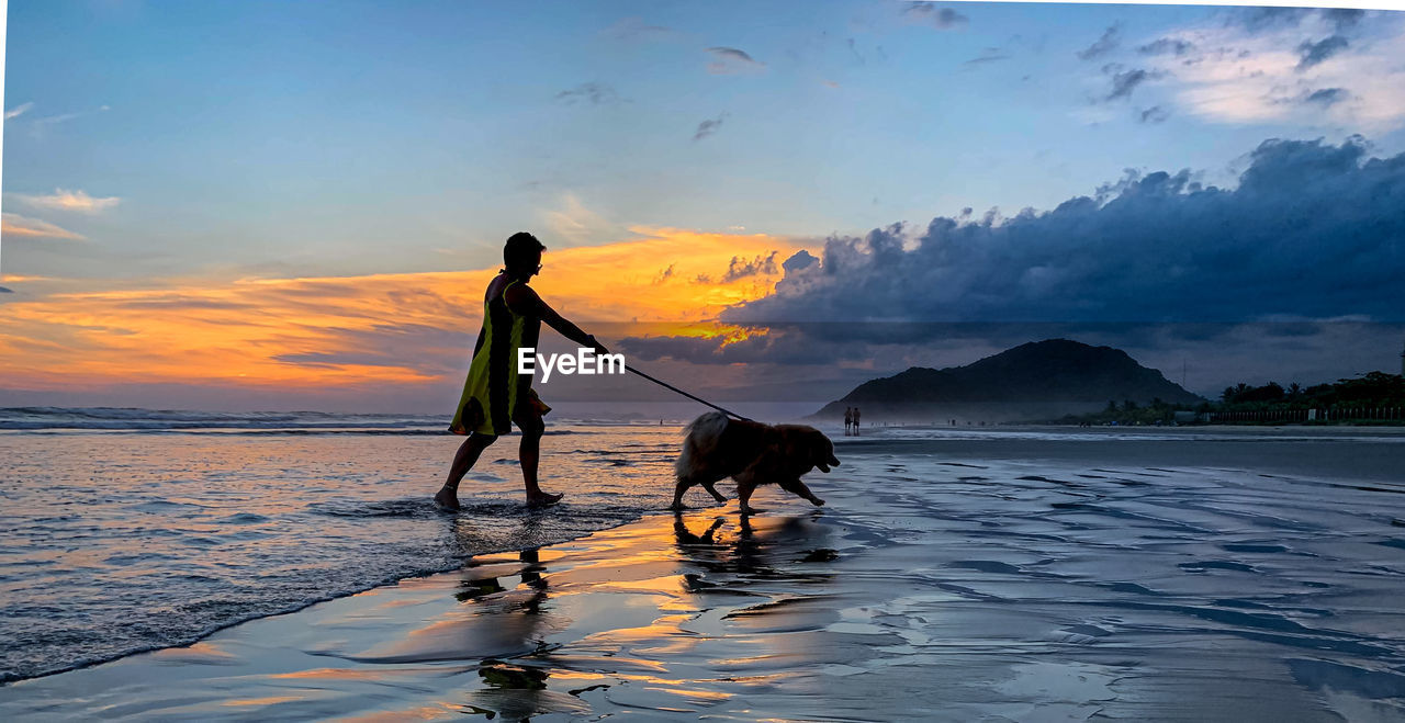 Woman with dog on beach against sky during sunset