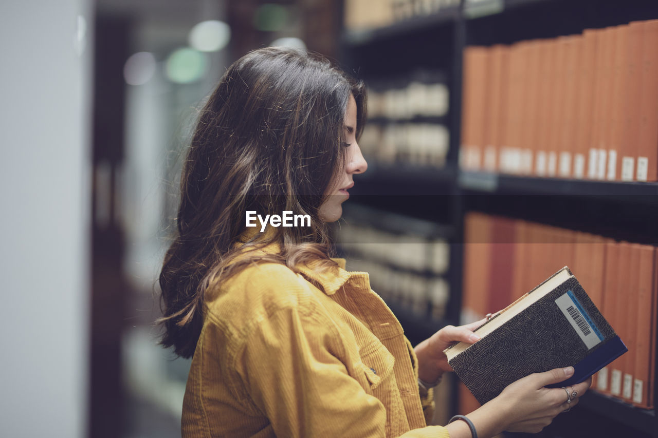 Side view of young woman holding book at library