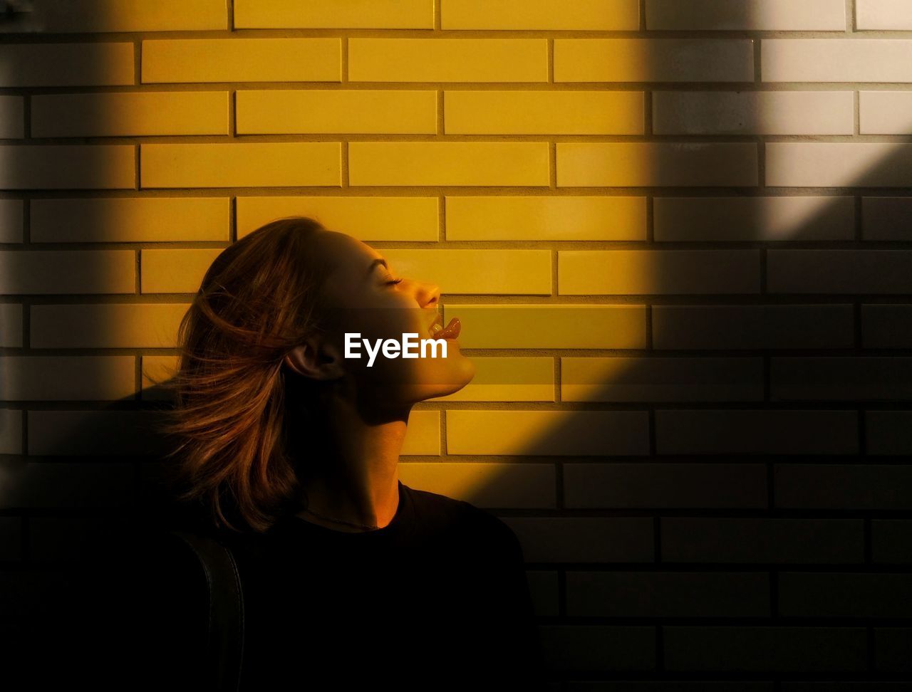 Side view of young woman sticking out tongue against wall during sunny day