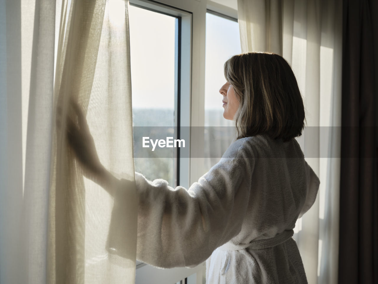 Blond senior woman opening white curtain while looking through window at luxury hotel room