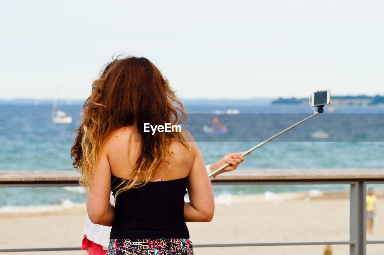 Rear view of friends taking selfie while standing by railing against sea