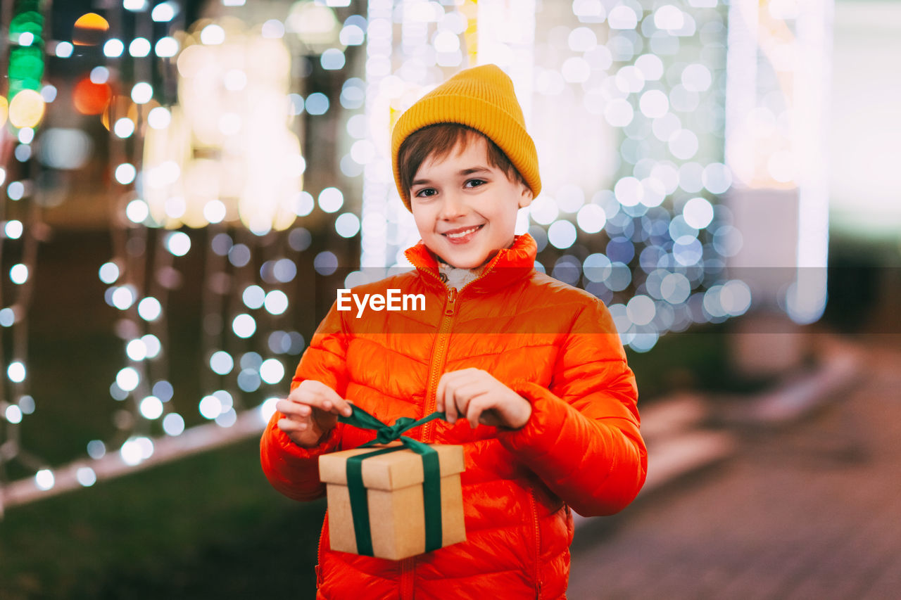 A happy boy in an orange jacket with a gift box in his hands at a christmas market on a winter 