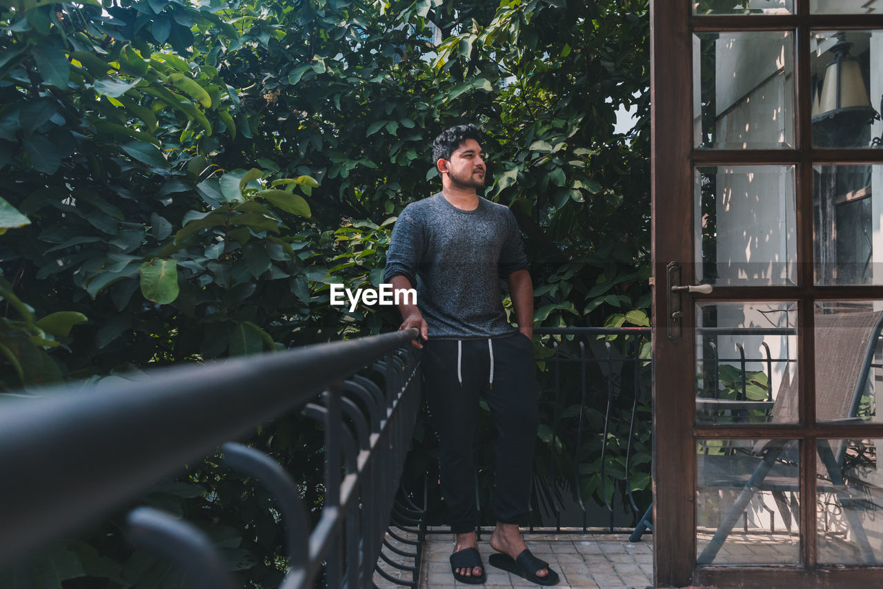 Portrait of young south asian man standing against trees while leaning on balcony railing 