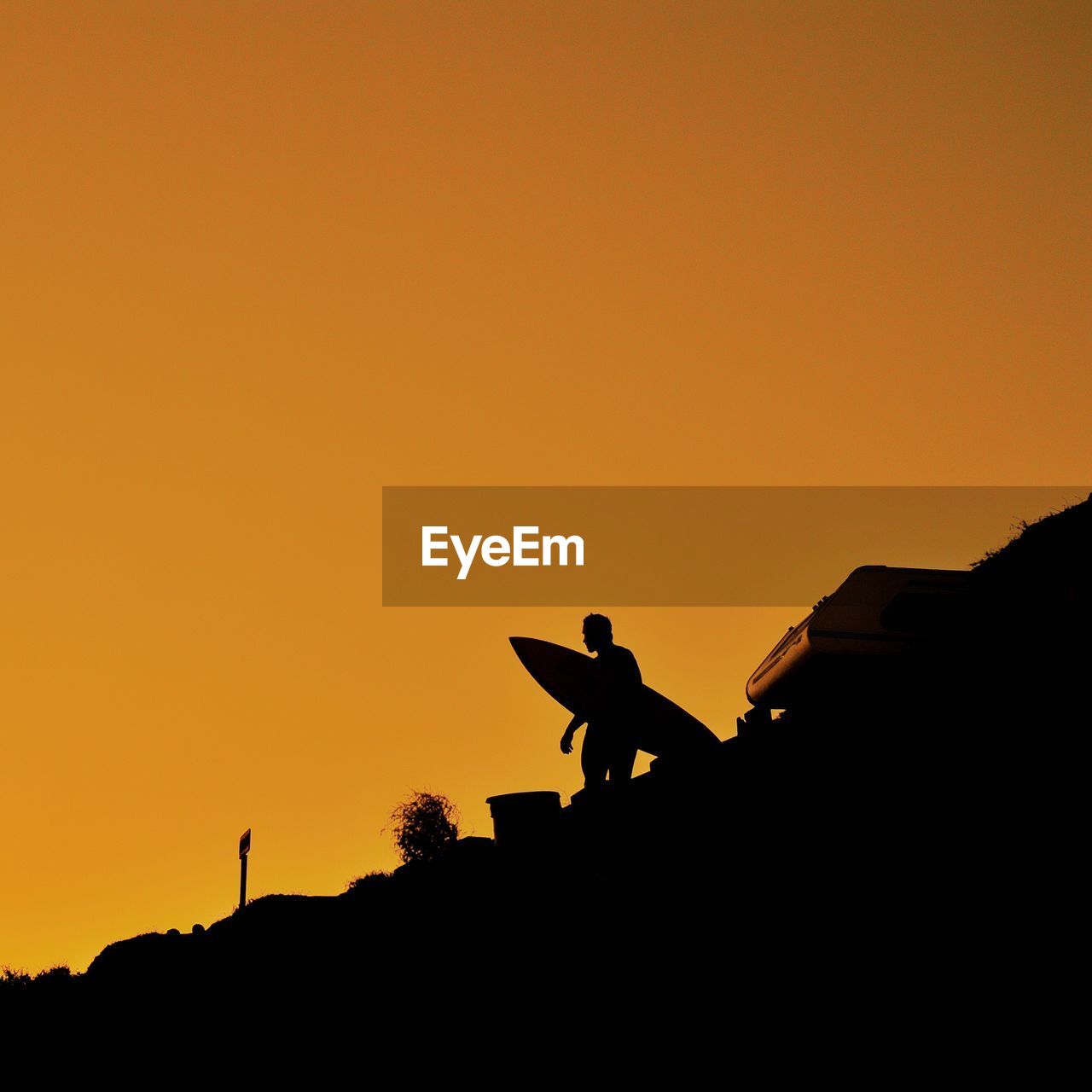 Low angle view of silhouette man with surfboard standing on land against orange sky