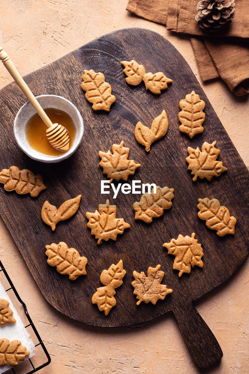 Top view abundance of leaf shaped sweet baked cookies placed on wooden cutting board near bowl of honey in kitchen