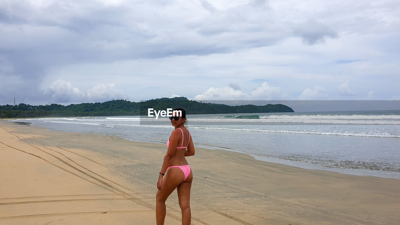 Portrait of smiling woman in bikini standing at beach against sky