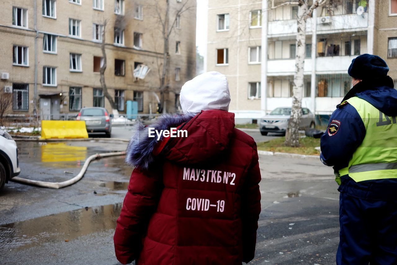 REAR VIEW OF PEOPLE STANDING ON STREET AMIDST BUILDINGS