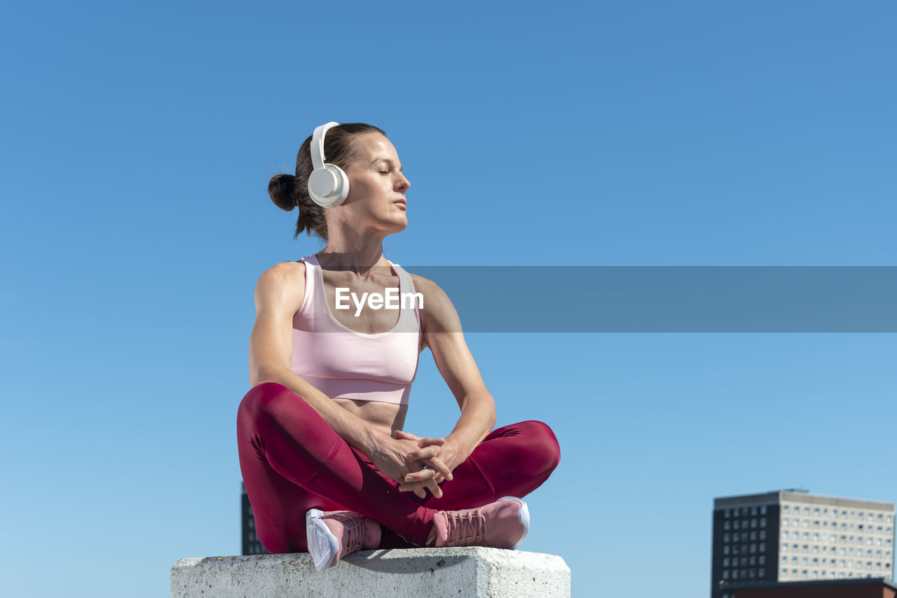 Attractive fit woman sitting crossed legged slistening to music with headphones outdoors in the sun.