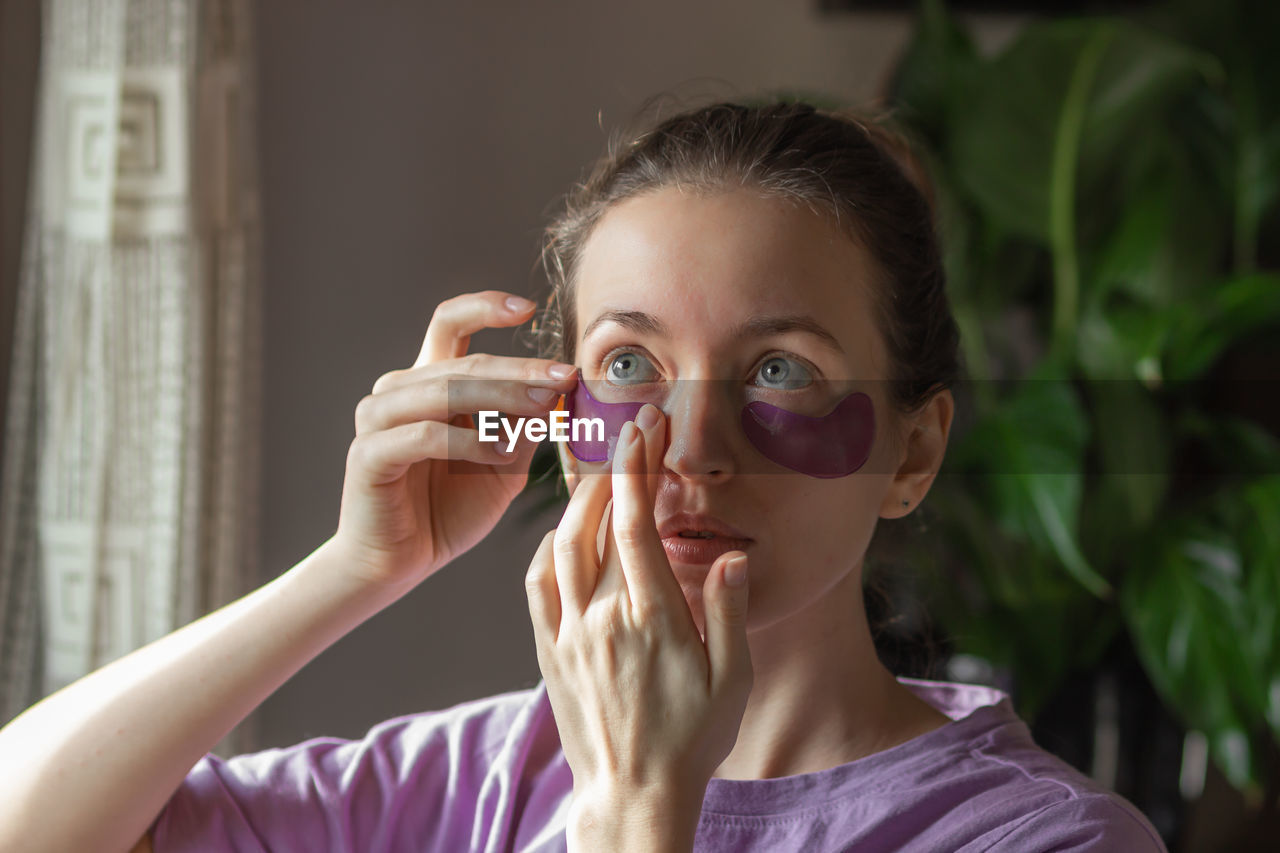 Young woman applying purple hydrogel eye patches against wrinkles, puffiness and dark circles.