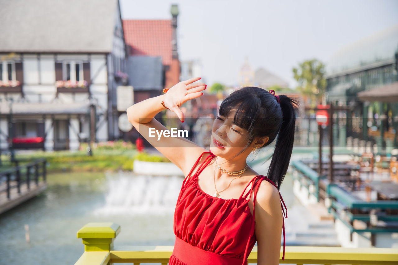 YOUNG WOMAN LOOKING AWAY WHILE STANDING ON CITY IN BACKGROUND