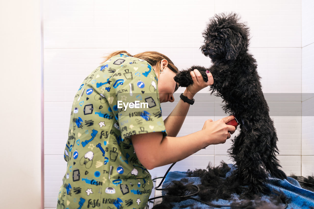 Woman groomer doing haircut with electric shaver for cute dog in clinic