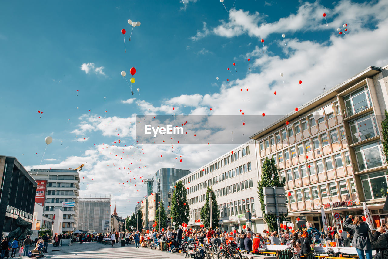 Low angle view of balloons flying over crowd and street amidst buildings on sunny day