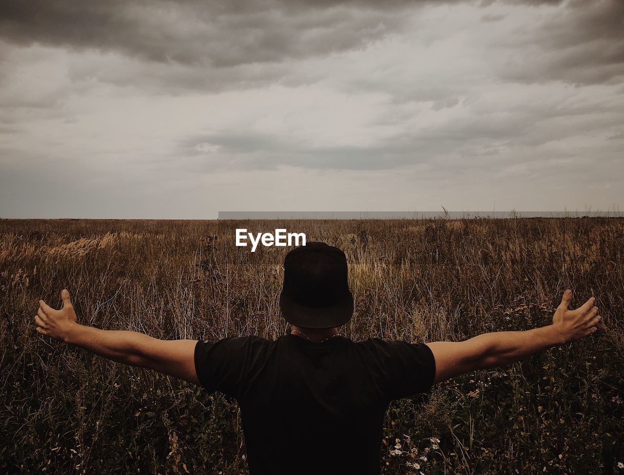 Man with arms outstretched on field against cloudy sky