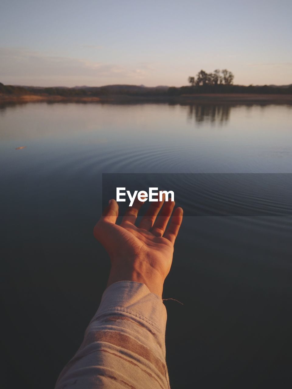 Cropped hand of person gesturing by lake against sky