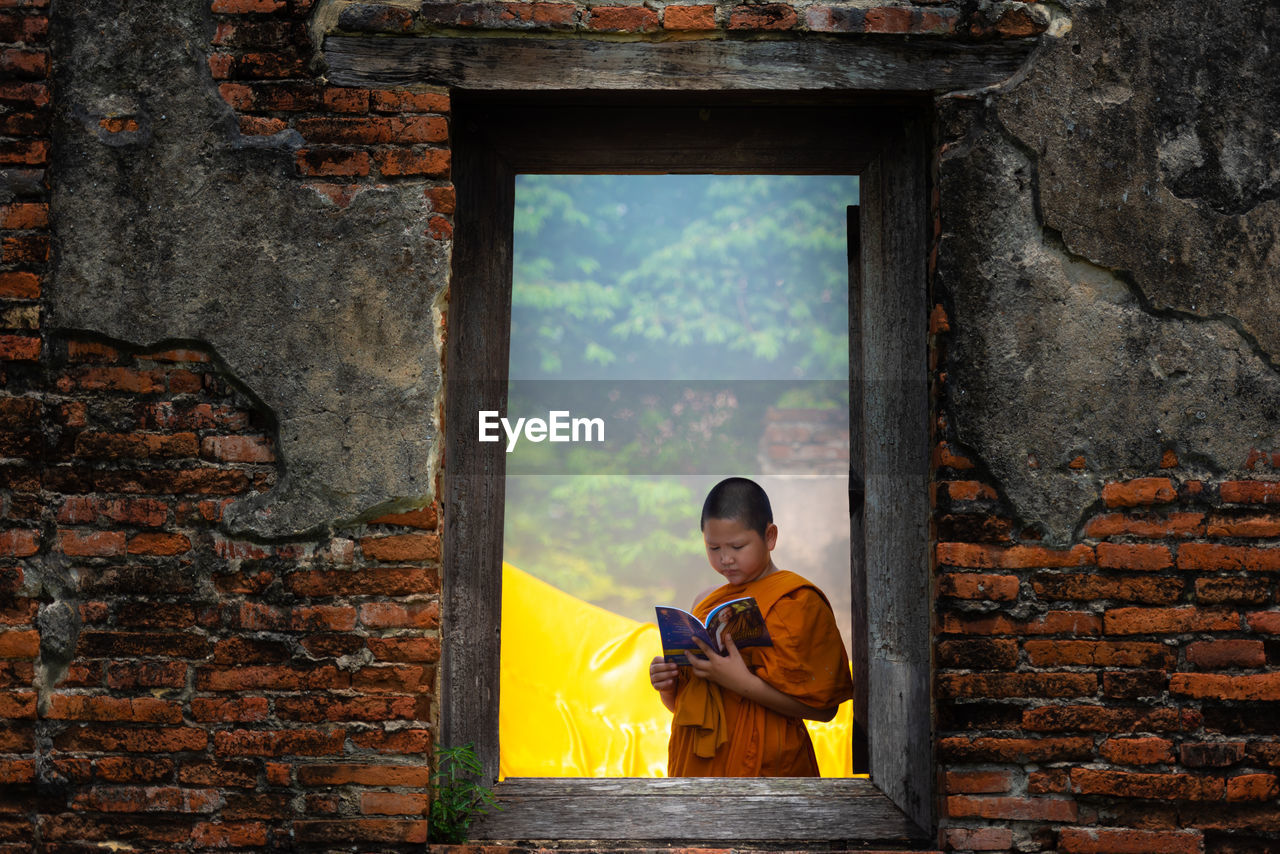 Boy wearing traditional clothing reading book seen through window