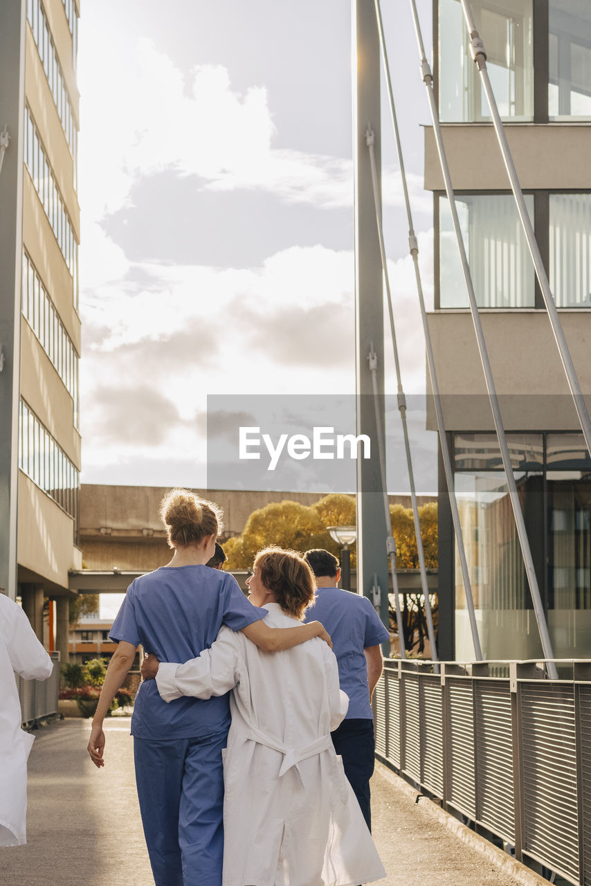 Rear view of healthcare workers with arms around each other walking towards hospital during sunset