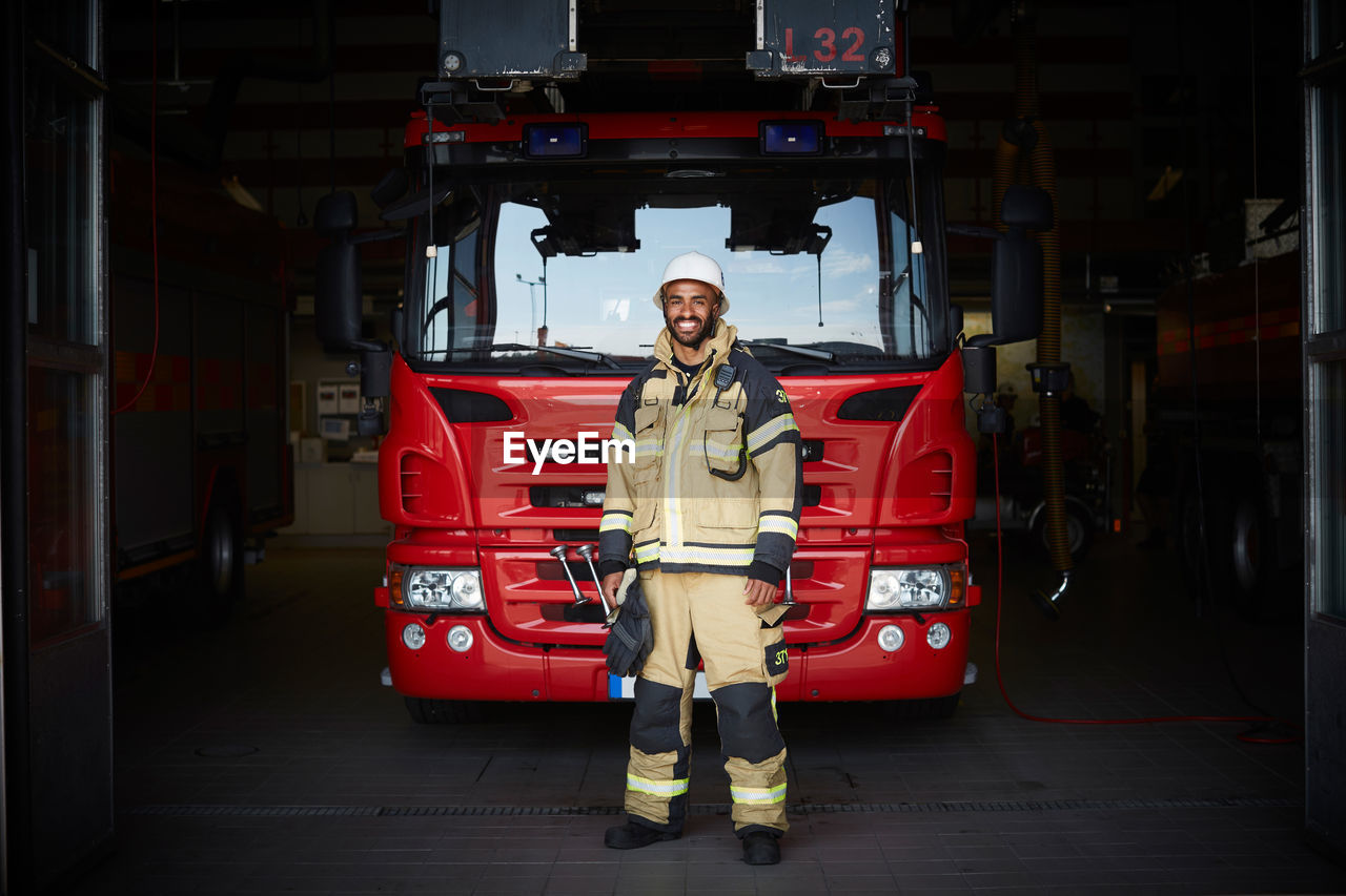 Full length portrait of smiling male firefighter standing in front of fire engine at fire station