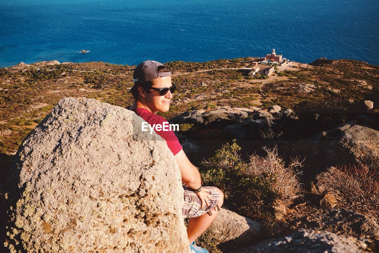 Young man wearing sunglasses sitting behind rock on hill against sea