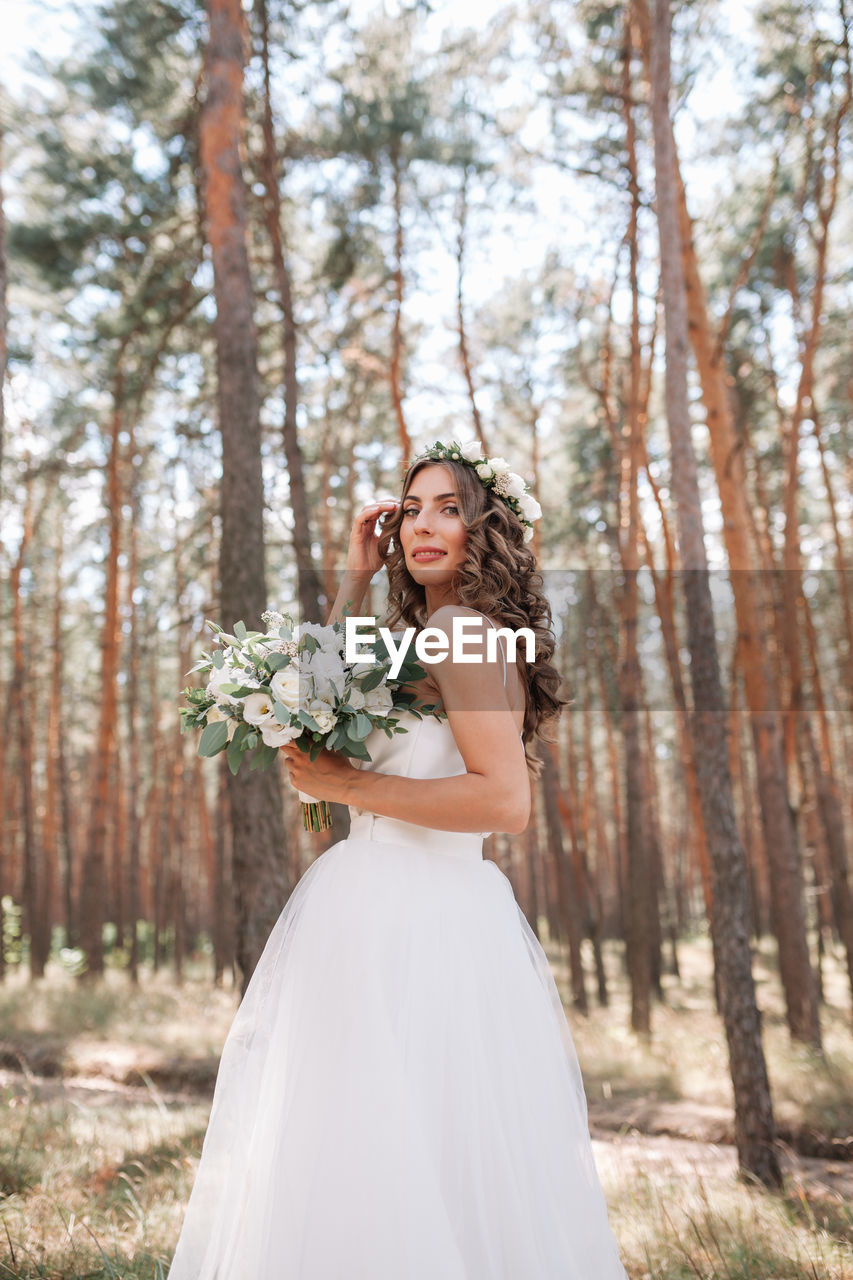 Portrait of young woman in wedding dress holding flower bouquet while standing against trees