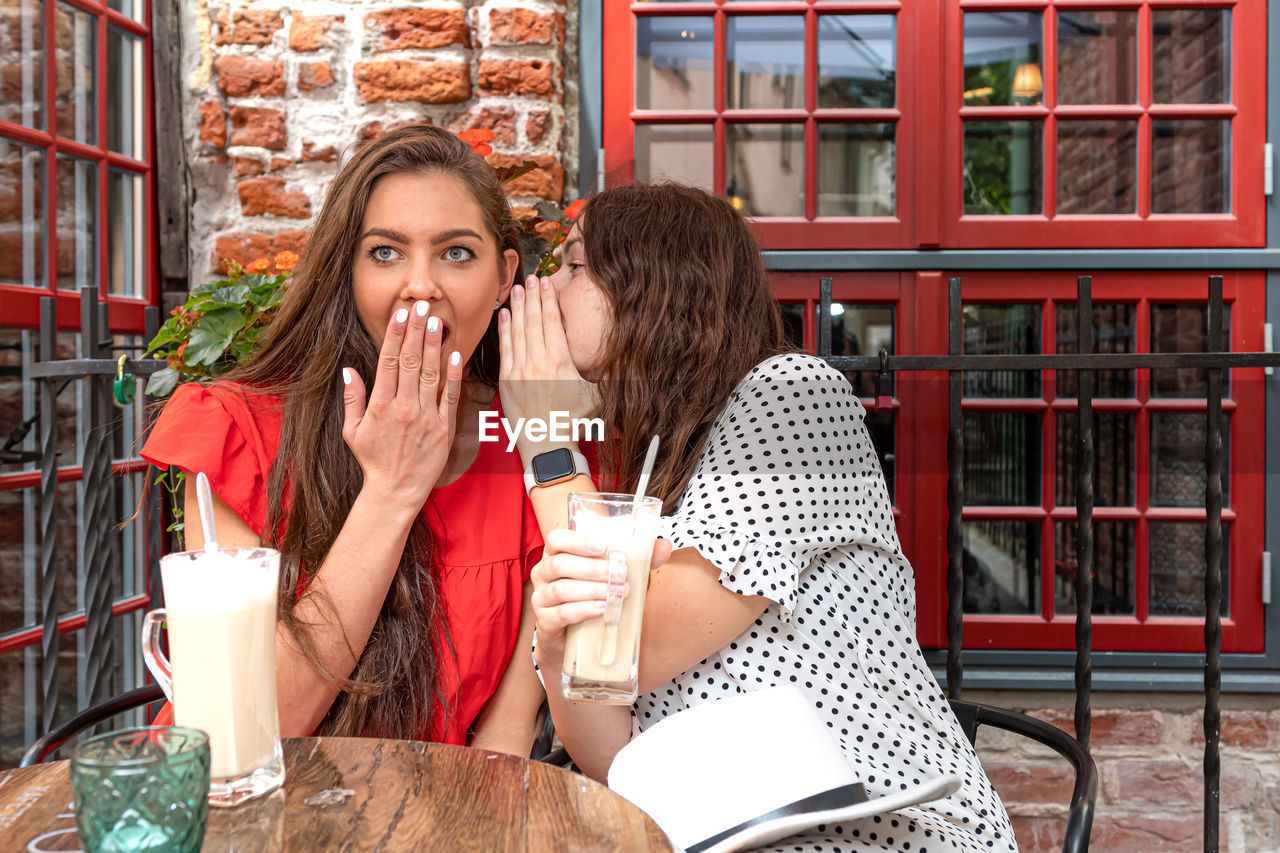 Two young beautiful girls sitting in urban cafe with coffee and sharing secrets, girls party concept