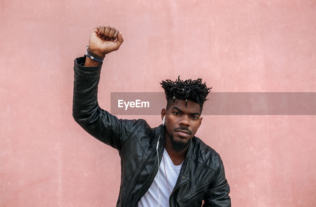 Serious man with arm raised listening music while standing against pink wall